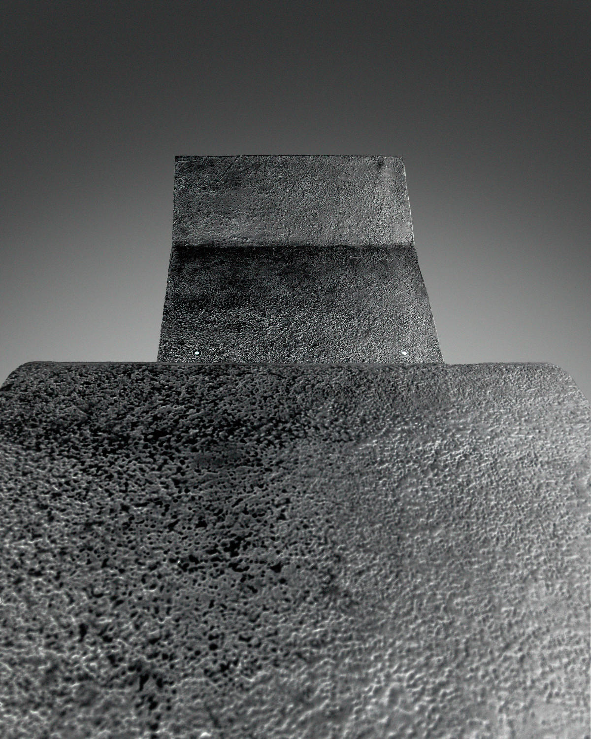 Close-up detail of a large dark textured metal slab sculpture shot from a low upward angle, revealing the rough granular surface and geometric form.