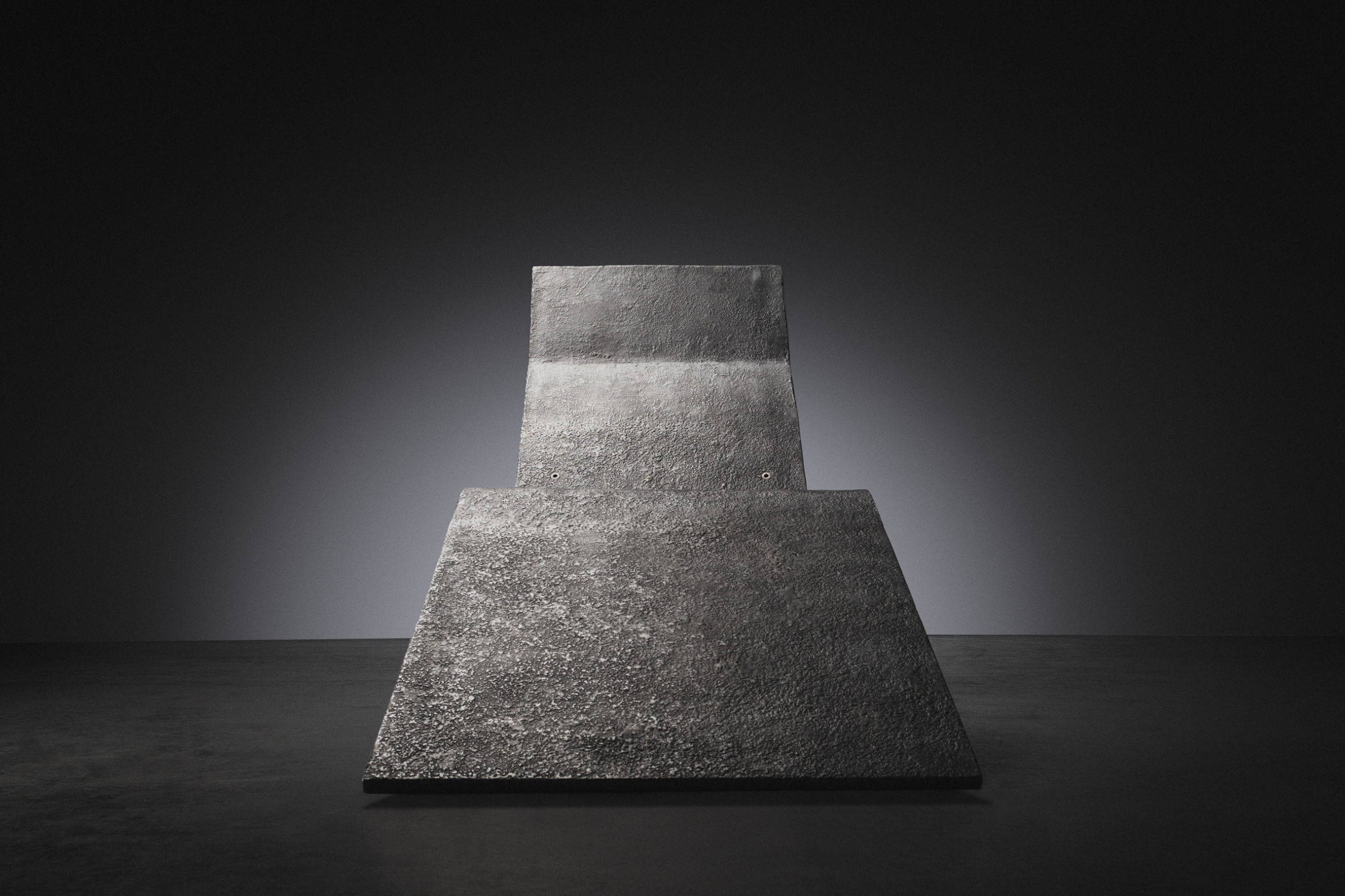 A large dark textured rectangular metal panel with a raw stone fragment at its base, set against a deep black gradient background.
