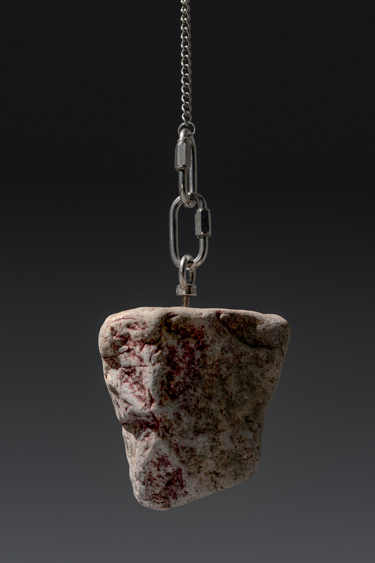 A rough pale stone with dark red mineral veining suspended mid-air by a steel chain and carabiner hardware against a dark gradient background.