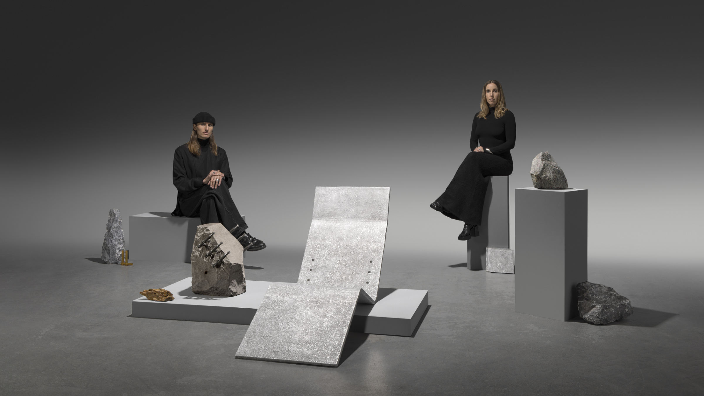 Batten & Kamp seated apart on white pedestals amid their sculptural works — a cast metal lounge chair, raw stones and mineral objects — dressed in black against a soft grey gradient.